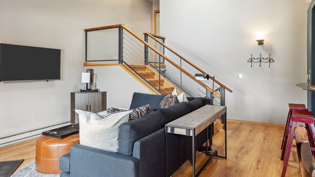 Un salon moderne avec un canapé, une table basse en bois, un téléviseur fixé au mur et un escalier doté d’une rampe en bois.