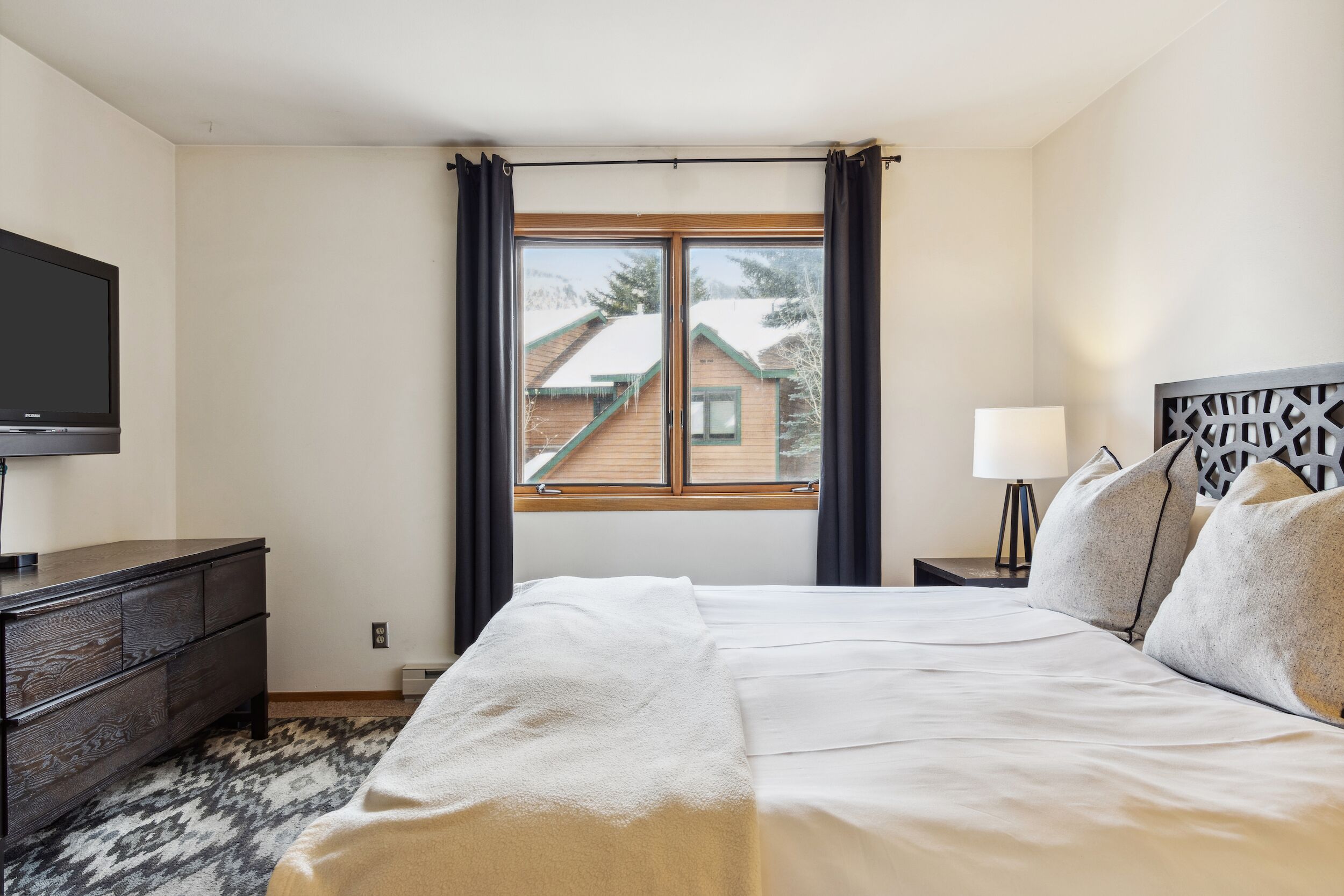Une chambre d’hôtel avec un lit, une commode, une télévision, une fenêtre avec des rideaux et une vue sur un bâtiment à l’extérieur.