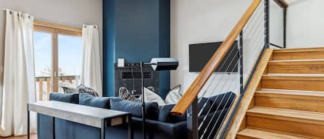 A modern living room with a wooden staircase, a sofa set, and a large window with curtains.