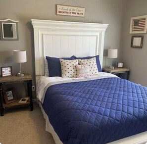 A bedroom with a white headboard, a blue quilted bedspread, and two nightstands with lamps. There are framed pictures on the nightstands and a sign on the wall that reads "Land of the Free BECAUSE OF THE BRAVE." - 3000 sq ft West Omaha home.  Perfect for wedding parties or families with kids! (Bennington)