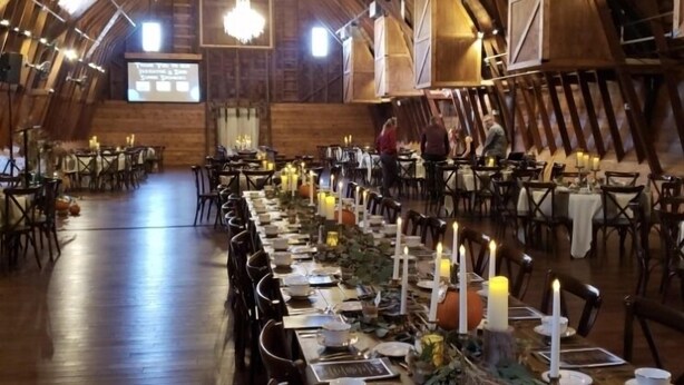 Una larga mesa de comedor preparada para una comida en un espacio rústico de estilo granero, con techos altos, vigas de madera y candelabros.