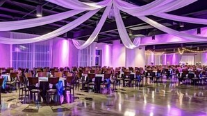 Un salón de banquetes con mesas dispuestas en filas, cubiertas con tela blanca e iluminado con luces moradas.