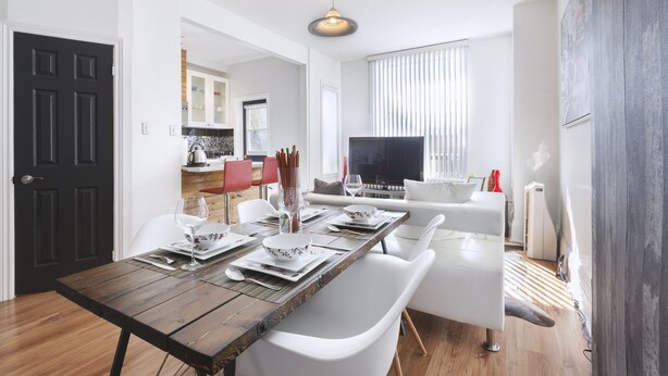 Un comedor moderno con una mesa de madera dispuesta para cuatro personas, sillas blancas, un televisor al fondo y una puerta negra a la izquierda.