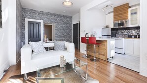 Una sala moderna con un sofá blanco, una mesa de centro de vidrio y un taburete rojo. Se observa una cocina con armarios blancos y una encimera de madera al fondo.