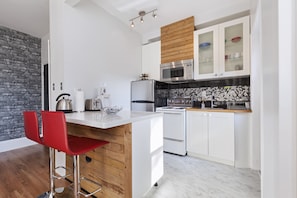 Una cocina moderna con armarios blancos, un muro decorativo de madera, un refrigerador de acero inoxidable, un microondas y una estufa. Hay dos taburetes rojos y una isla blanca con un lavamanos.
