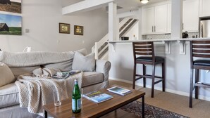 A living room with a sofa, coffee table, and a view of a kitchen with white cabinets and a staircase. - Outpost: Balsam Lodge A6 (Wilson)