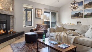 A modern living room with a fireplace, leather sofa, and a coffee table with books and a bottle. - Outpost: Balsam Lodge A6 (Wilson)