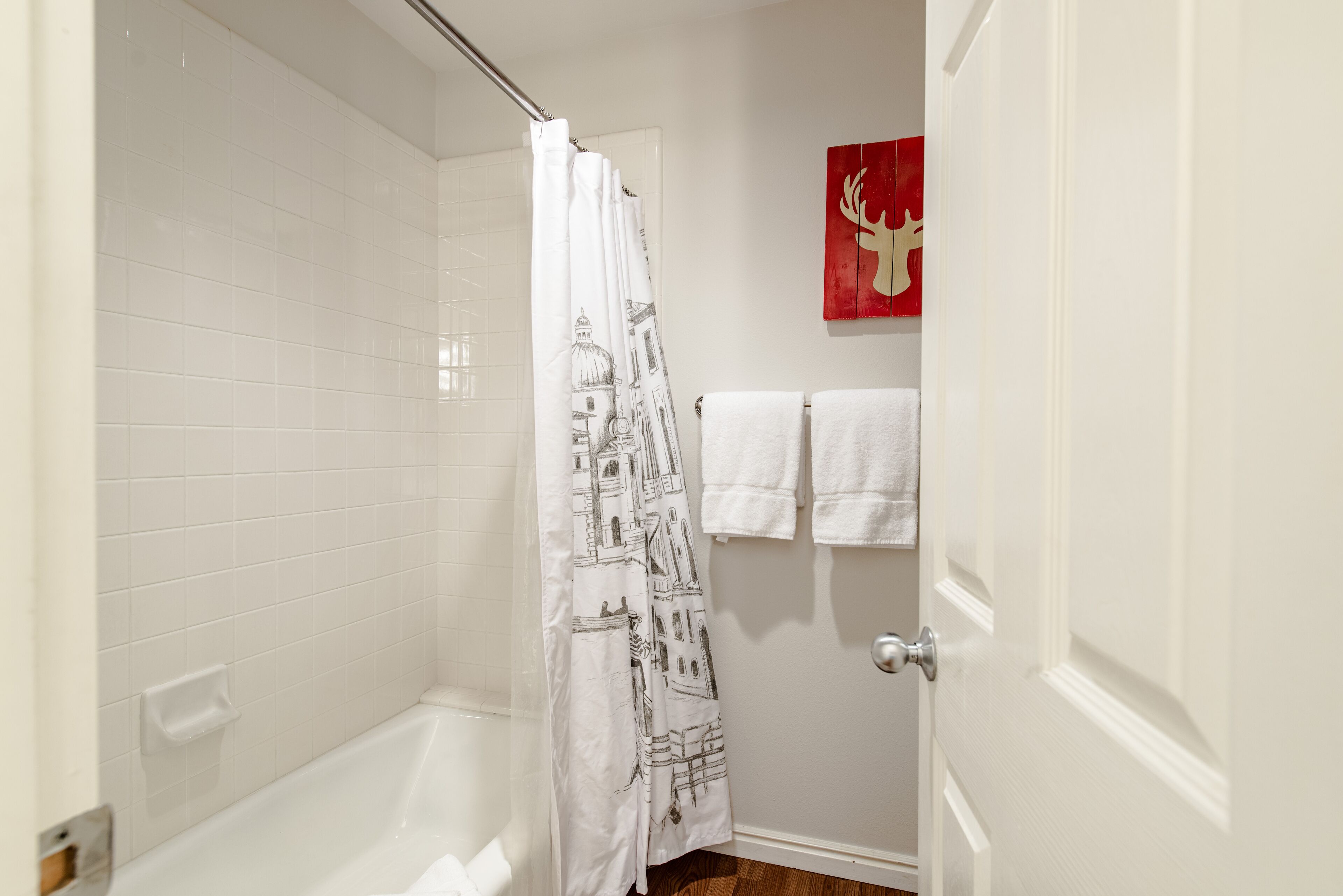 A bathroom with a shower curtain featuring architectural prints, a white bathtub, and a door with a red artwork of a deer.