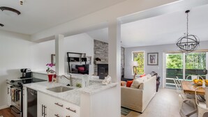 A modern kitchen with white cabinetry, a built-in oven, and a dishwasher. There is a dining area with a wooden table and chairs, and a living room with a sofa and a coffee table. - Outpost: Balsam Lodge A6 (Wilson)