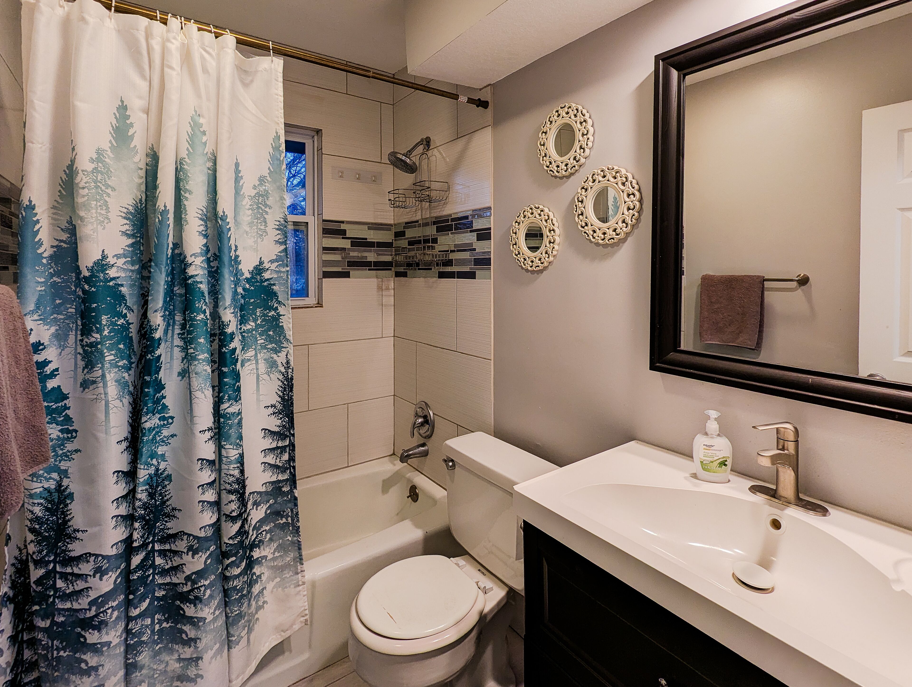 A bathroom with a shower curtain featuring a forest design, a toilet, a sink with a mirror above it, and decorative wall mirrors.