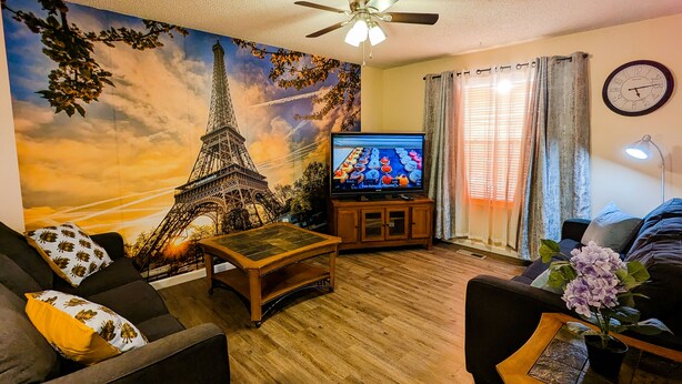 A living room with a large mural of the Eiffel Tower, a television, a ceiling fan, and a clock on the wall. - Spacious and comfortable 3 bedrooms house in the heart of Overland Park (Overland Park)