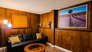 A room with wooden panel walls, a black leather sofa with yellow cushions, a wooden coffee table, a floor lamp, and a large window with a lavender field view. - Spacious and comfortable 3 bedrooms house in the heart of Overland Park (Overland Park)
