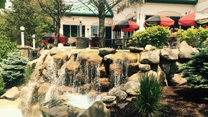 A waterfall feature with surrounding rocks and a small pond, a tree, and a building with outdoor seating in the background. -  Lake Life Luxury, Lake Geneva, Short Walk To Downtown, Pool View Upgraded Suite (Lake Geneva)