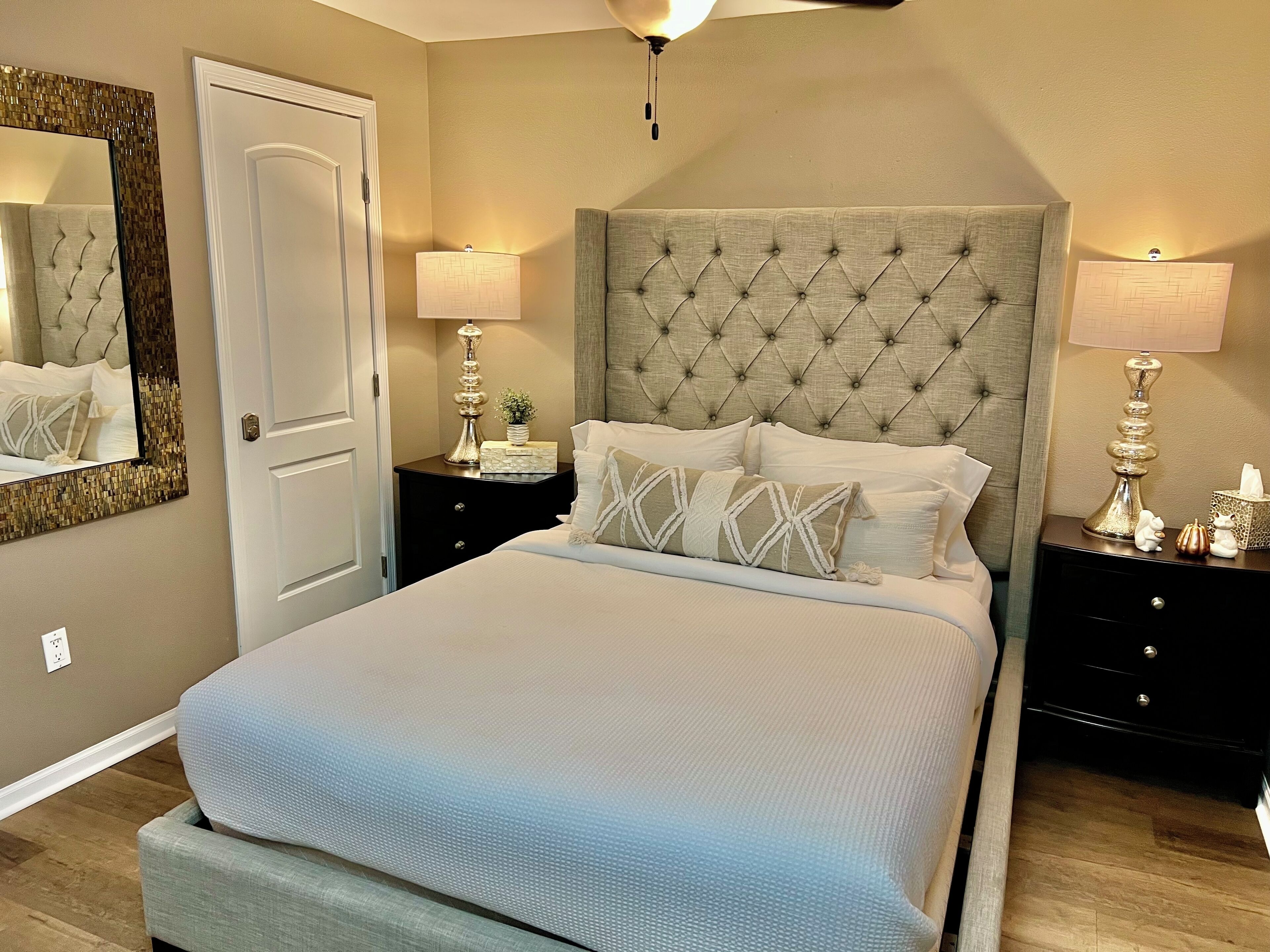 Une chambre à coucher avec une tête de lit capitonnée, un grand lit recouvert d’une couverture bleu clair, deux tables de chevet avec des lampes et un miroir au mur.