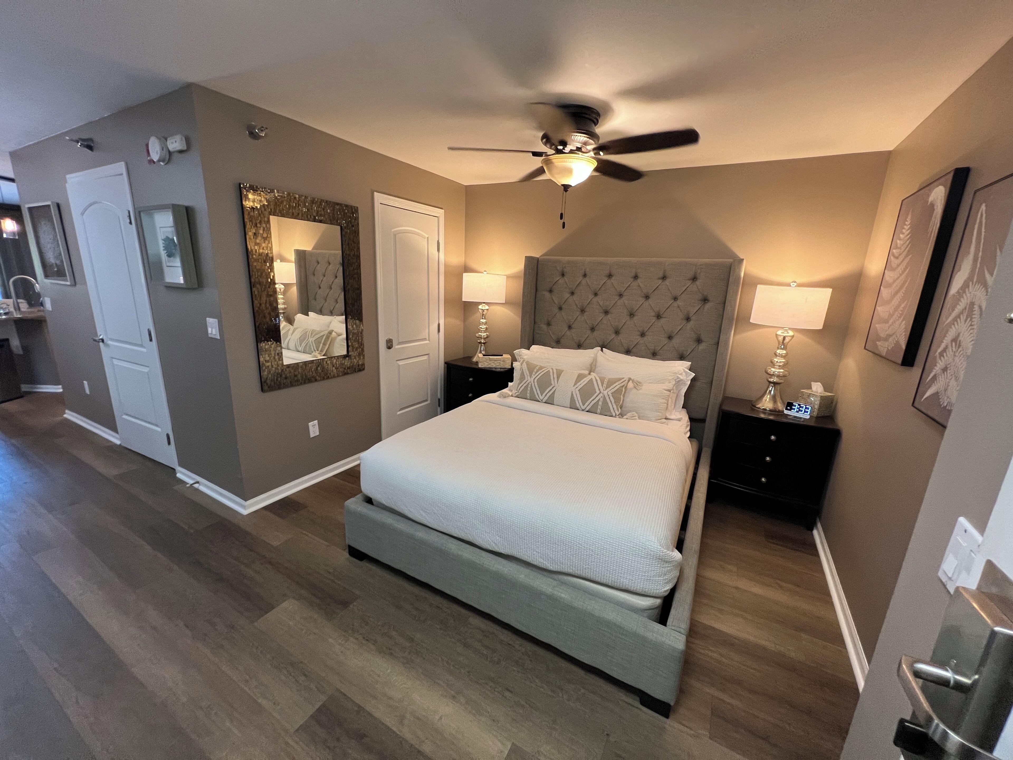 A bedroom with a large bed, a ceiling fan, two bedside tables with lamps, a mirror, and a door.