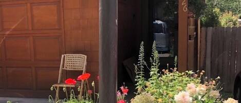 Un edificio de madera con puerta de garaje, una silla de madera y varias plantas y flores en primer plano.