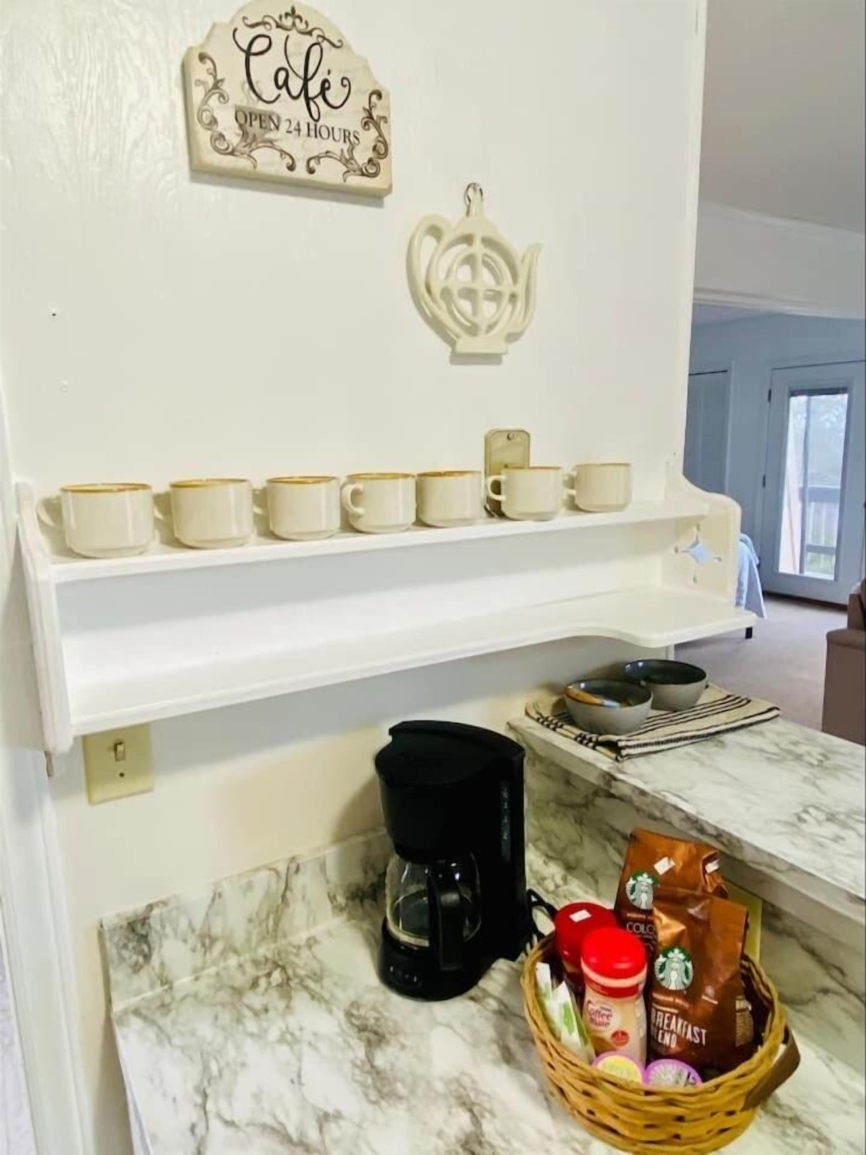 A kitchen with a coffee station, including a coffee maker, cups, and a basket with coffee bags.