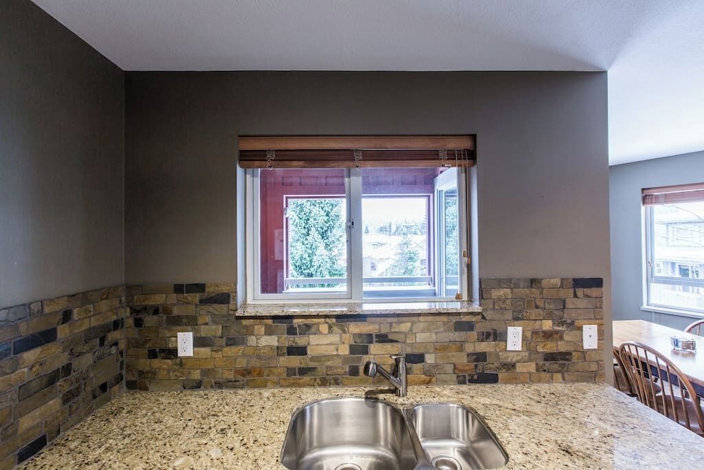 A kitchen with granite countertops, a double sink, a window with a view, and a stone backsplash.