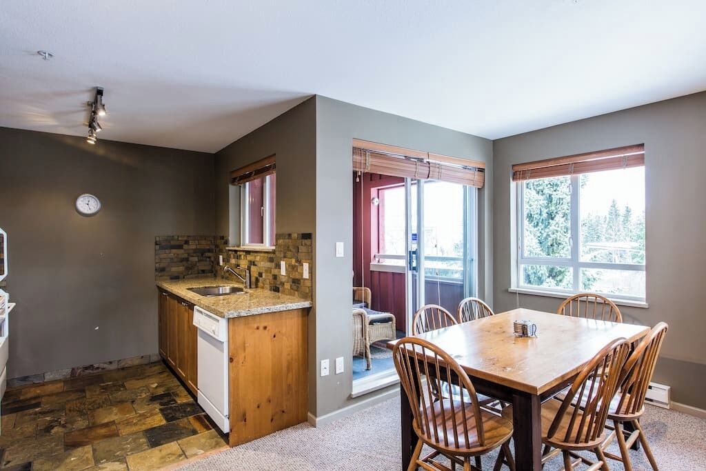A kitchen with a granite countertop, wooden cabinets, and a dishwasher. There is a dining area with a wooden table and chairs.