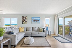 A living room with a grey sectional sofa, a round pouf, a wooden coffee table, and a view of the ocean through large windows.
