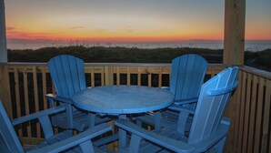 Une terrasse en bois avec une table ronde et deux chaises bleues, donnant sur un coucher de soleil sur la plage.