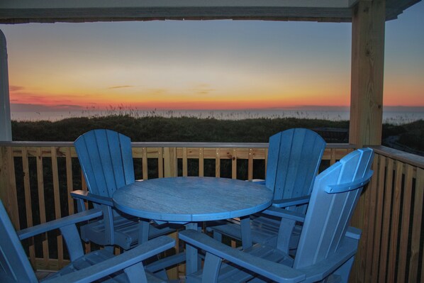 Una terraza de madera con una mesa redonda y dos sillas azules mirando hacia un atardecer sobre la playa.
