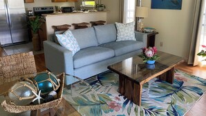 A living room with a grey sofa, a glass coffee table, and a floral rug. There is a kitchen in the background with wooden cabinets and a refrigerator. - 1BR/1BA BEACHFRONT • GREAT LOCATION • July 2026-Sept 2026 Discounted! (Kihei)