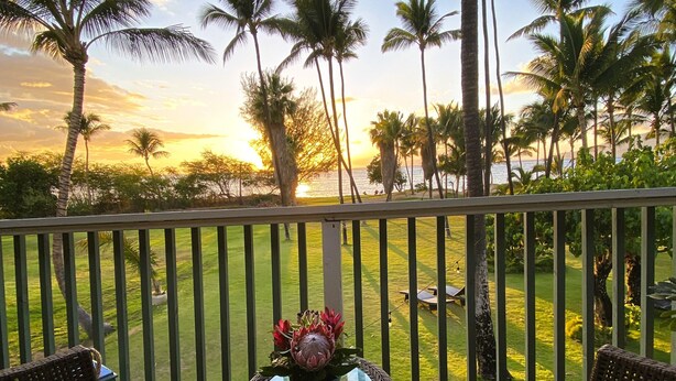 A balcony with wicker furniture and a table with a flower arrangement, overlooking a grassy area with palm trees and a sunset view. - 1BR/1BA BEACHFRONT • GREAT LOCATION • July 2026-Sept 2026 Discounted! (Kihei)