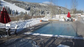 A heated outdoor pool with lounge chairs and umbrellas, surrounded by snow-covered terrain and mountains.