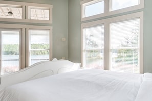 A bedroom with a large bed, white headboard, and multiple windows with blinds.