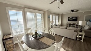 A dining area with a wooden table and white chairs, a ceiling fan, and large windows with blinds.