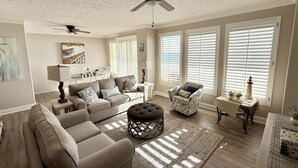 A spacious living room with a large sofa, a coffee table, and a ceiling fan. There are large windows with blinds, a dining area with a table and chairs, and a small side table with a lamp.