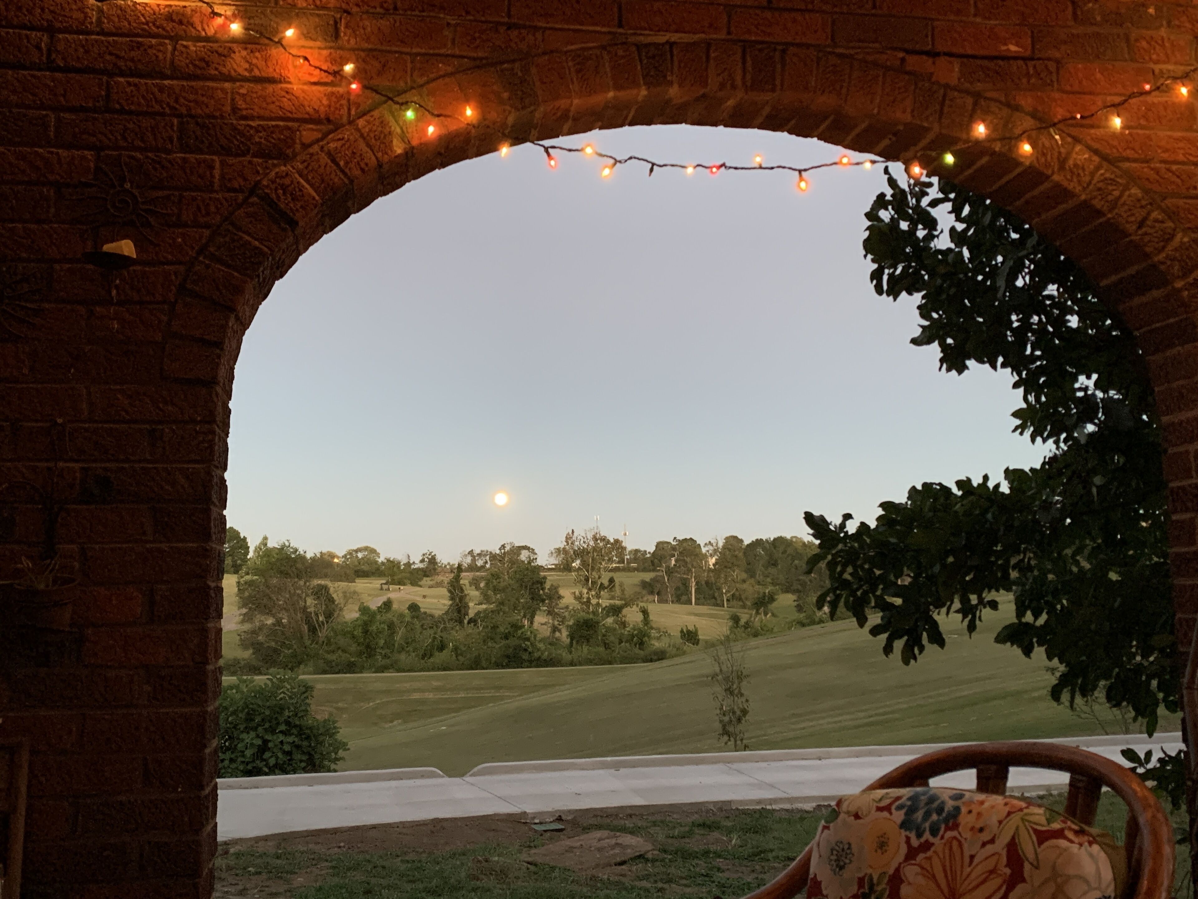 A brick archway with string lights, a view of a green landscape, and a chair with a floral cushion.