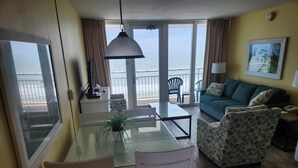 A living room with a glass table, white chairs, a blue sofa, and a view of the ocean.