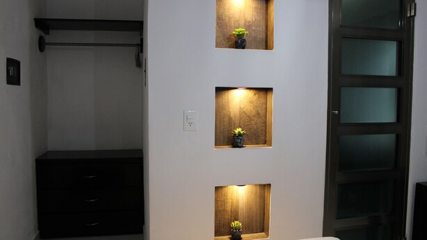 Quarto com cama, cômoda e três prateleiras fixadas na parede, iluminadas e com pequenas plantas em vasos.