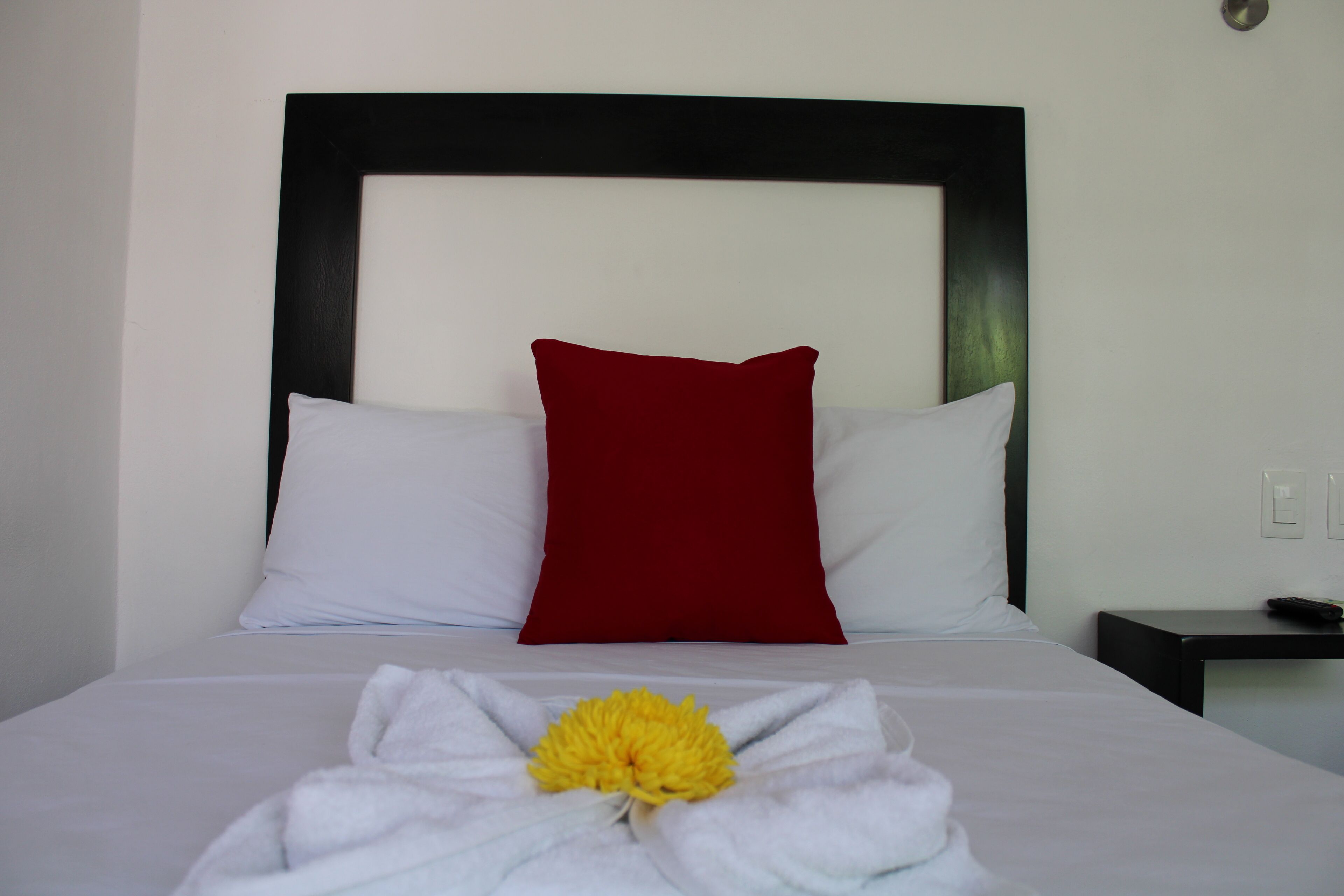 Una cama bien hecha con almohadas blancas y rojas, un edredón blanco y una flor amarilla sobre la colcha.
