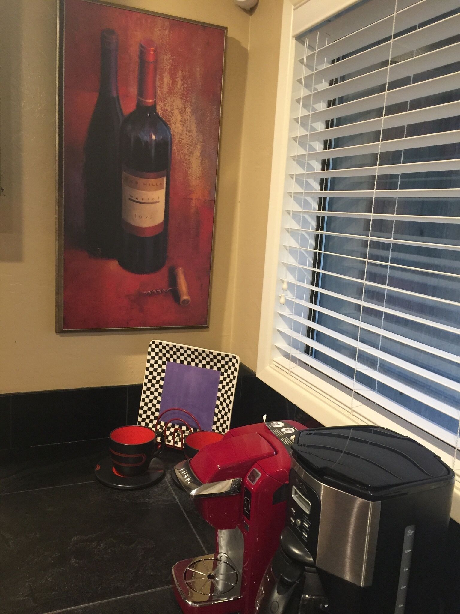 Une cuisine équipée d’une machine à café, d’une chaise à carreaux, d’un tableau représentant une bouteille de vin et d’une fenêtre avec des stores.