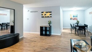 A modern living room with a black sofa, a small side table with a plant, a dining area with a table and chairs, and a large mirror on the wall. - Cute and affordable 3BDR duplex in Overland Park for up to 12 people! (Overland Park)