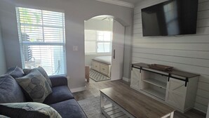 A living room with a sofa, coffee table, TV, and kitchen area with cabinets and a window.