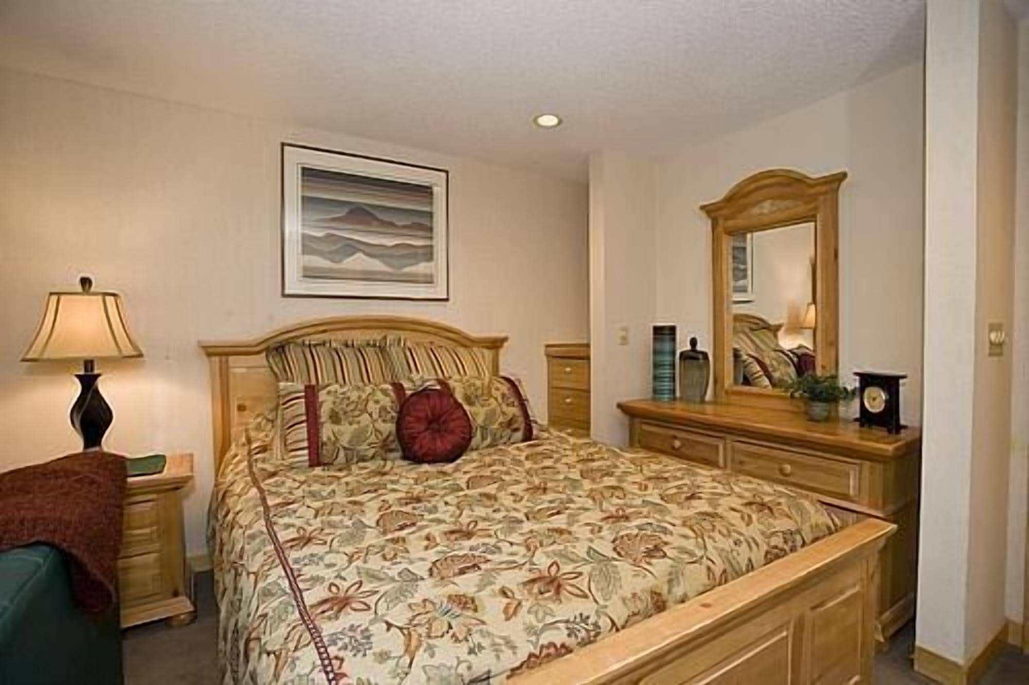 Un dormitorio con cama, mesita de noche, cómoda, espejo y un cuadro en la pared.
