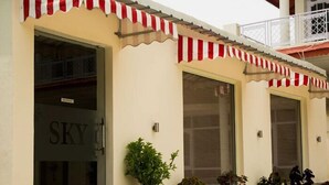 Un bâtiment à la façade beige, doté d’auvents rayés rouges et blancs, et de nombreux pots de plantes placés devant l’entrée.