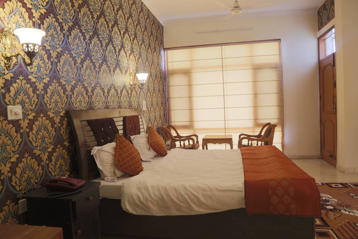 Quarto de hotel com uma cama grande, duas cadeiras, uma mesinha e papel de parede estampado.