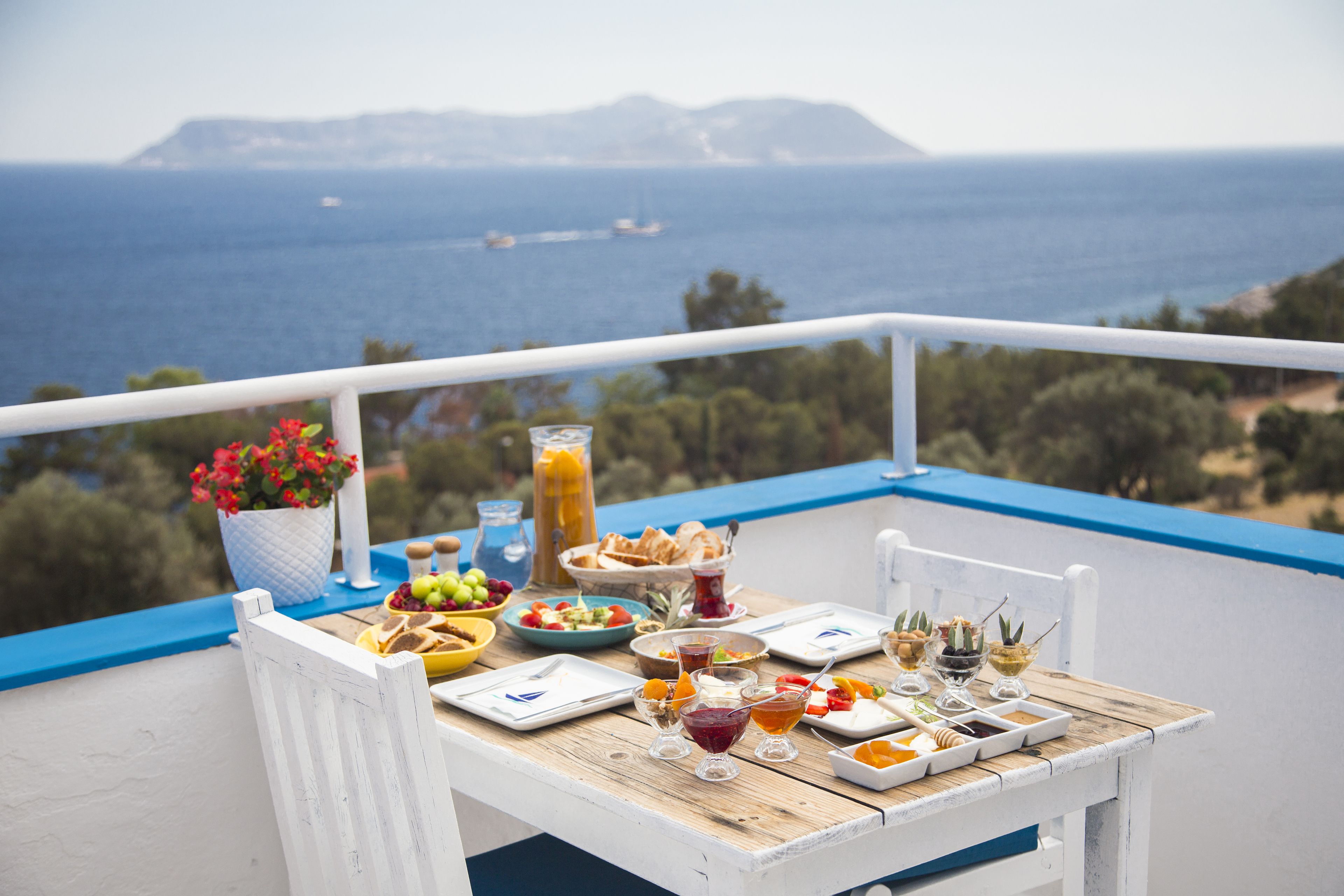 A wooden table with a variety of food and drinks on a balcony overlooking the sea.