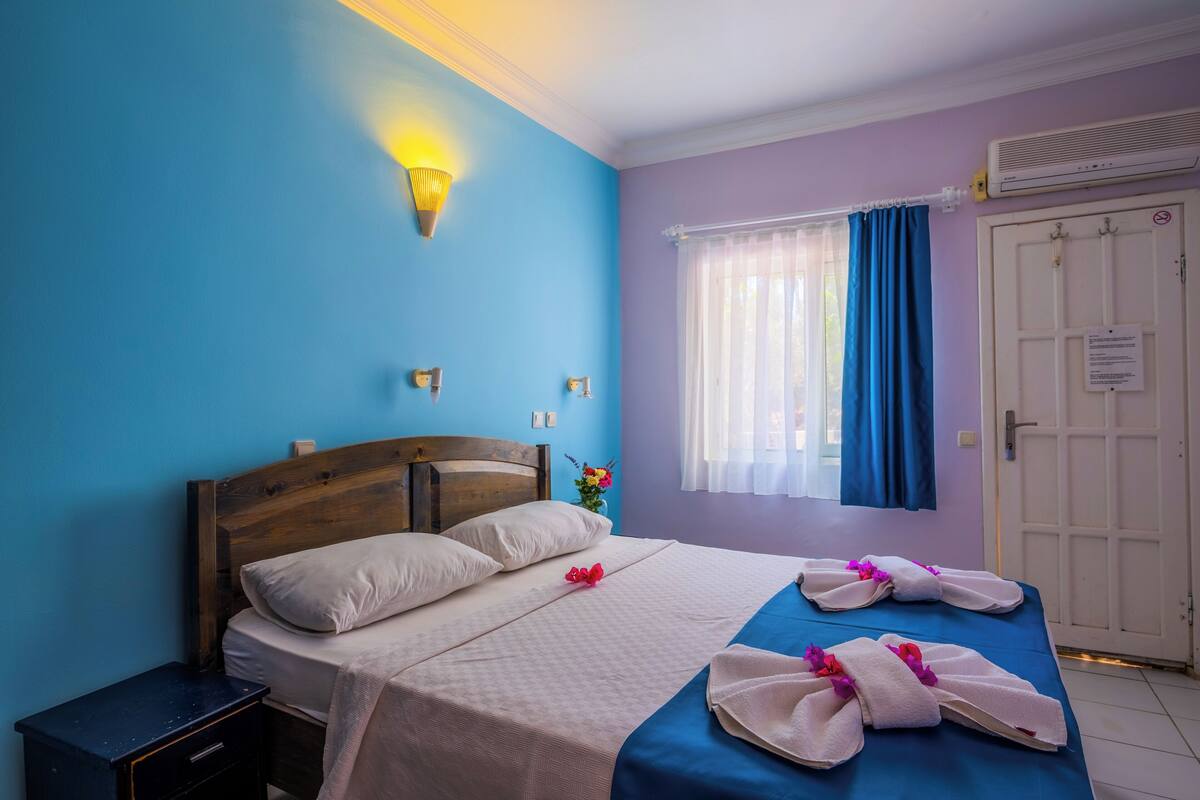 A bedroom with a bed, a nightstand, a wall-mounted light, a window with curtains, and a door with a sign.