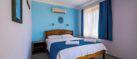Quarto com cama de madeira, mesa de cabeceira, ar condicionado fixado na parede e janela com cortinas azuis.