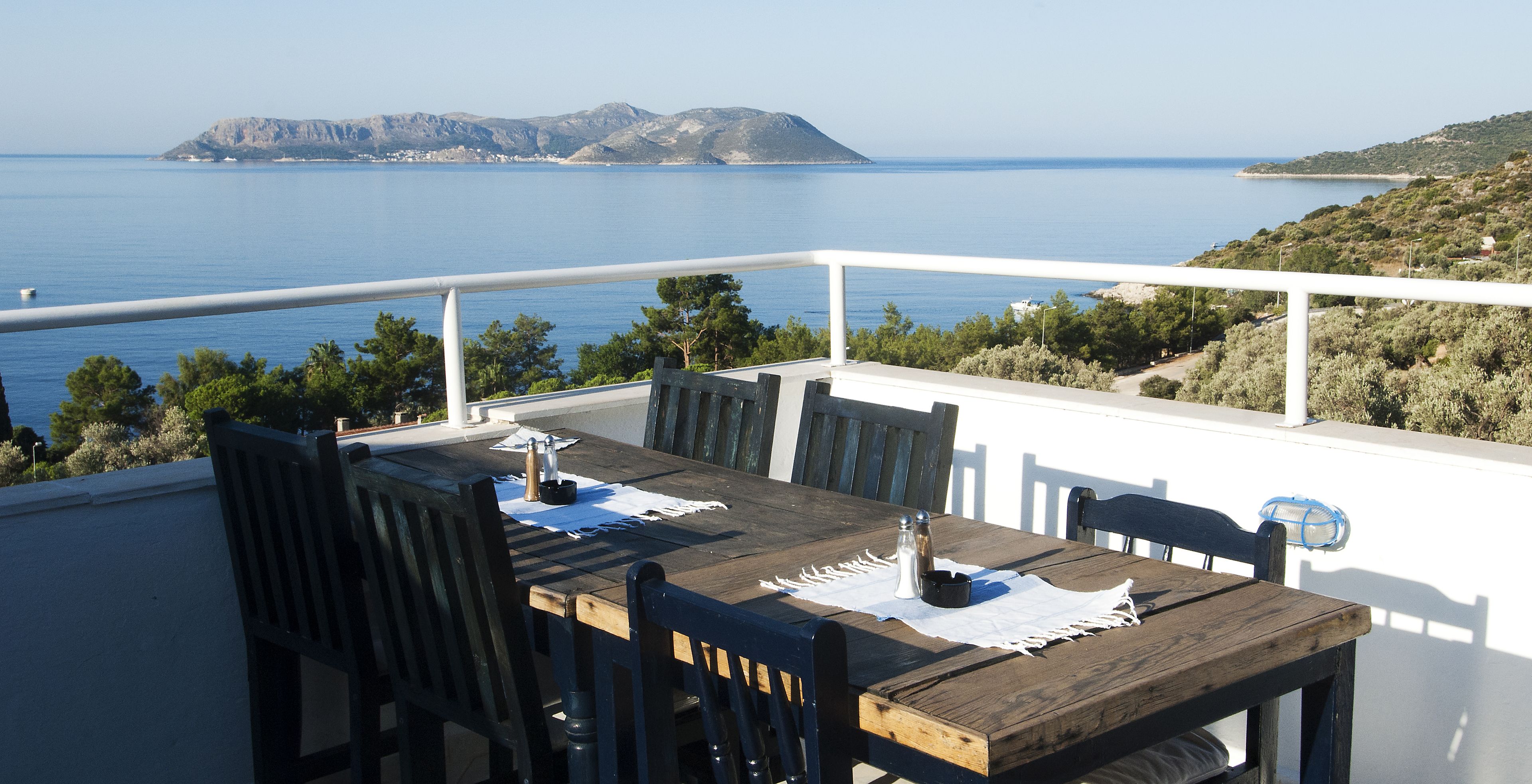 A wooden table with chairs and a white railing overlooking a body of water and hills.