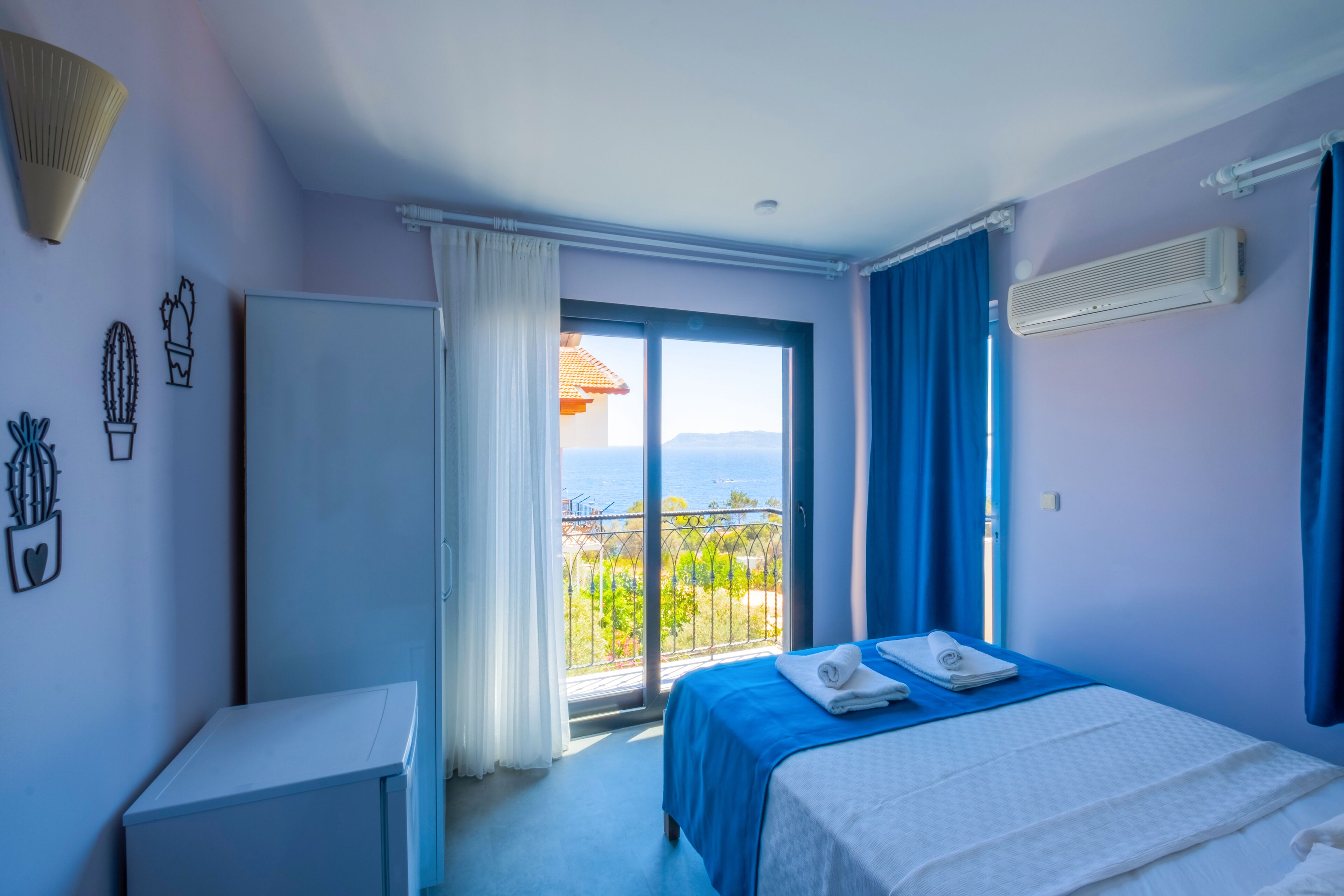 Ein Schlafzimmer mit Balkon, einem Bett mit blauer Bettdecke, einem weißen Nachttisch und einer Wand-Klimaanlage.