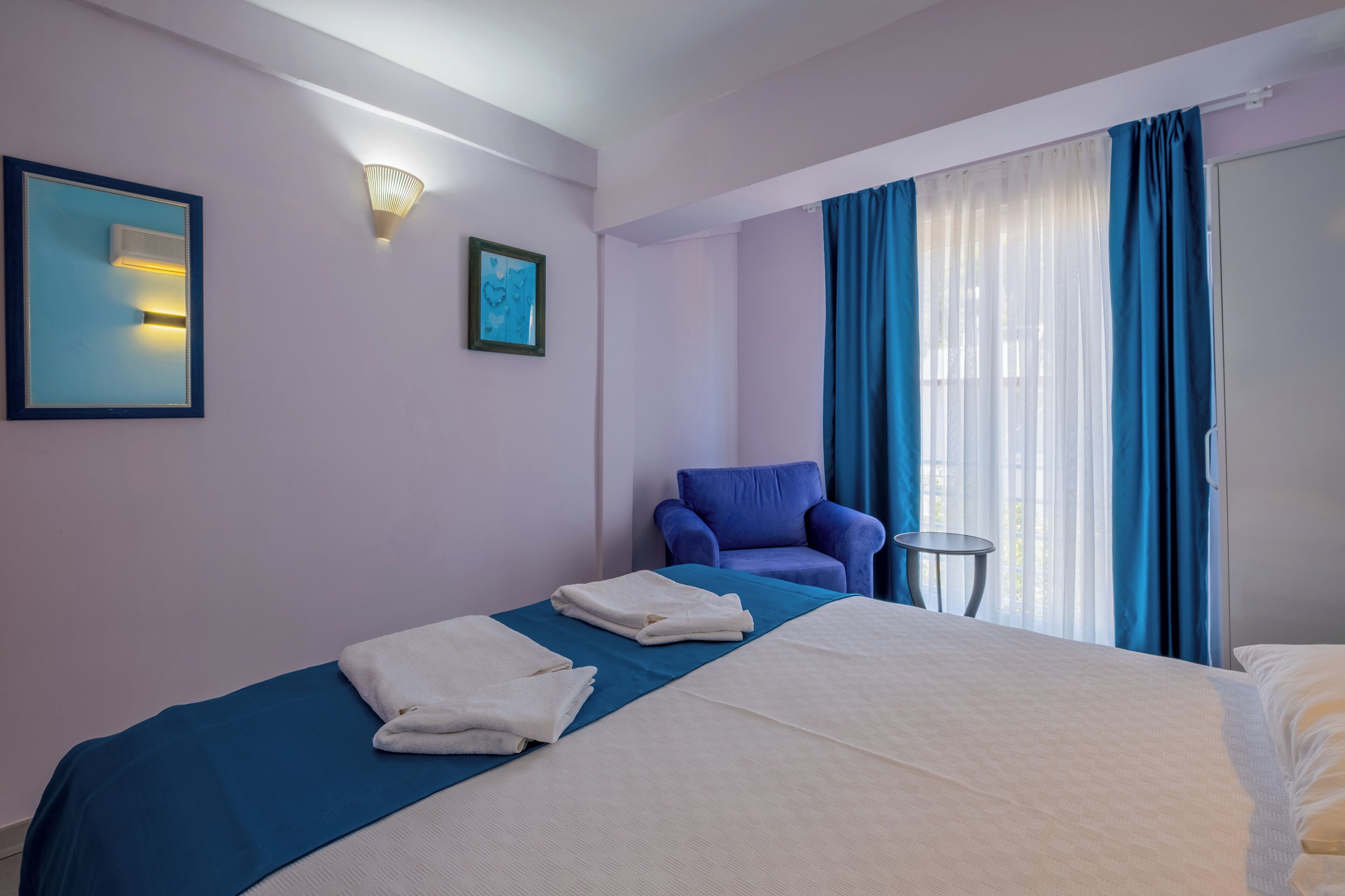 A hotel room with a bed, a chair, a small table, and a window with curtains.