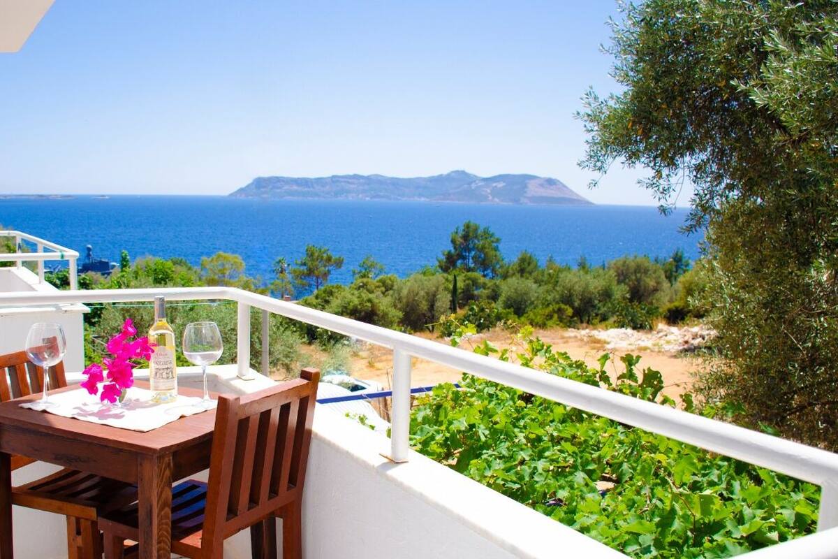 A balcony with a table set for two, overlooking a coastal landscape with clear blue sea and distant hills.