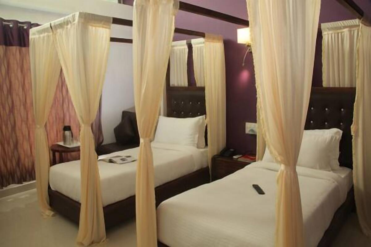 Quarto de hotel com duas camas, cada uma com dossel. Há uma mesa de cabeceira com uma lâmpada e um telefone em uma das camas.
